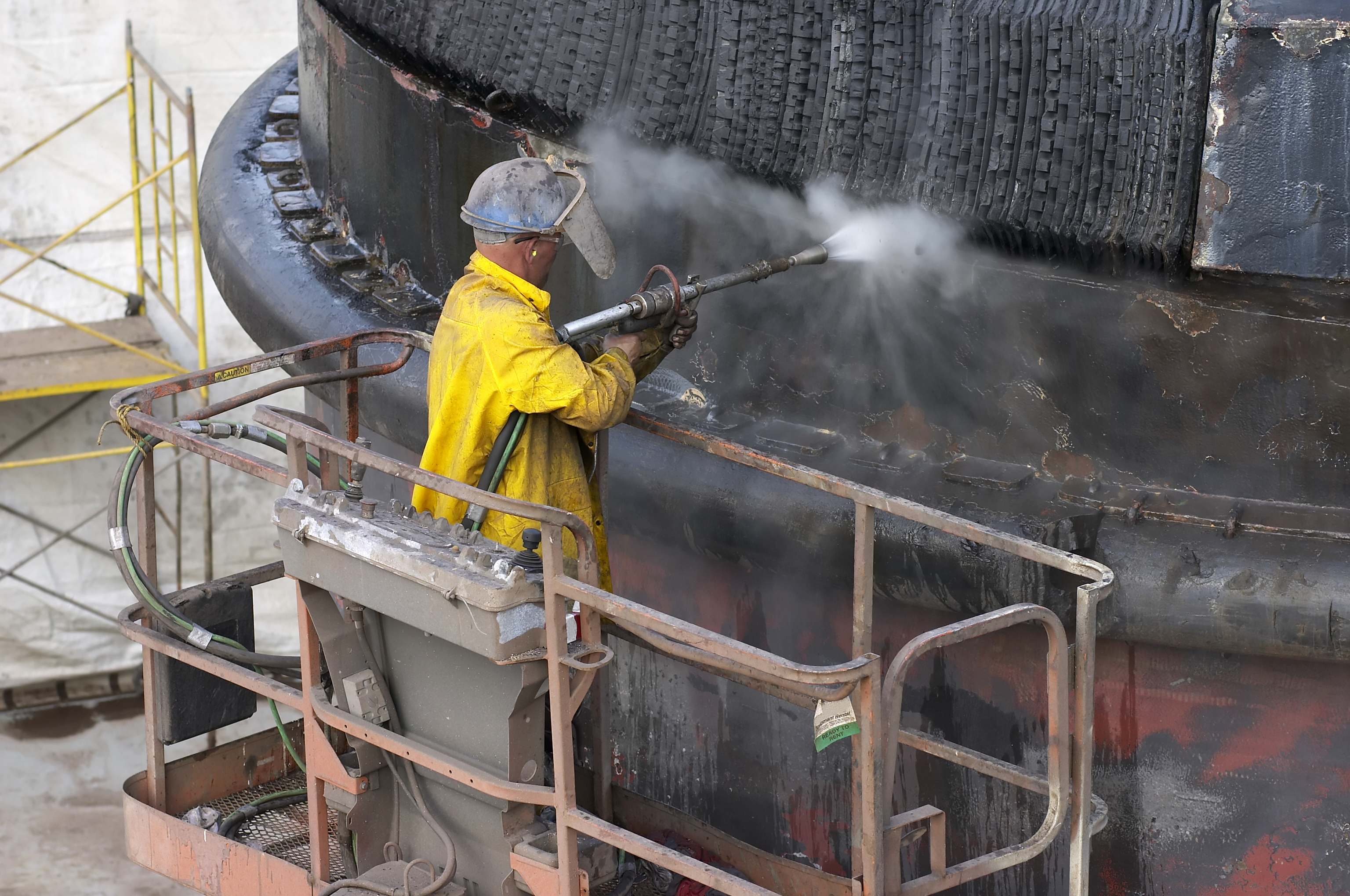 A man in the centre of the image is wearing a dark blue, metal helmet and a yellow, long-sleeved jacket. He is facing away and is holding a dark grey blasting machine. He's blasting water onto a large, cylinder structure which has a dark, flaking coating.