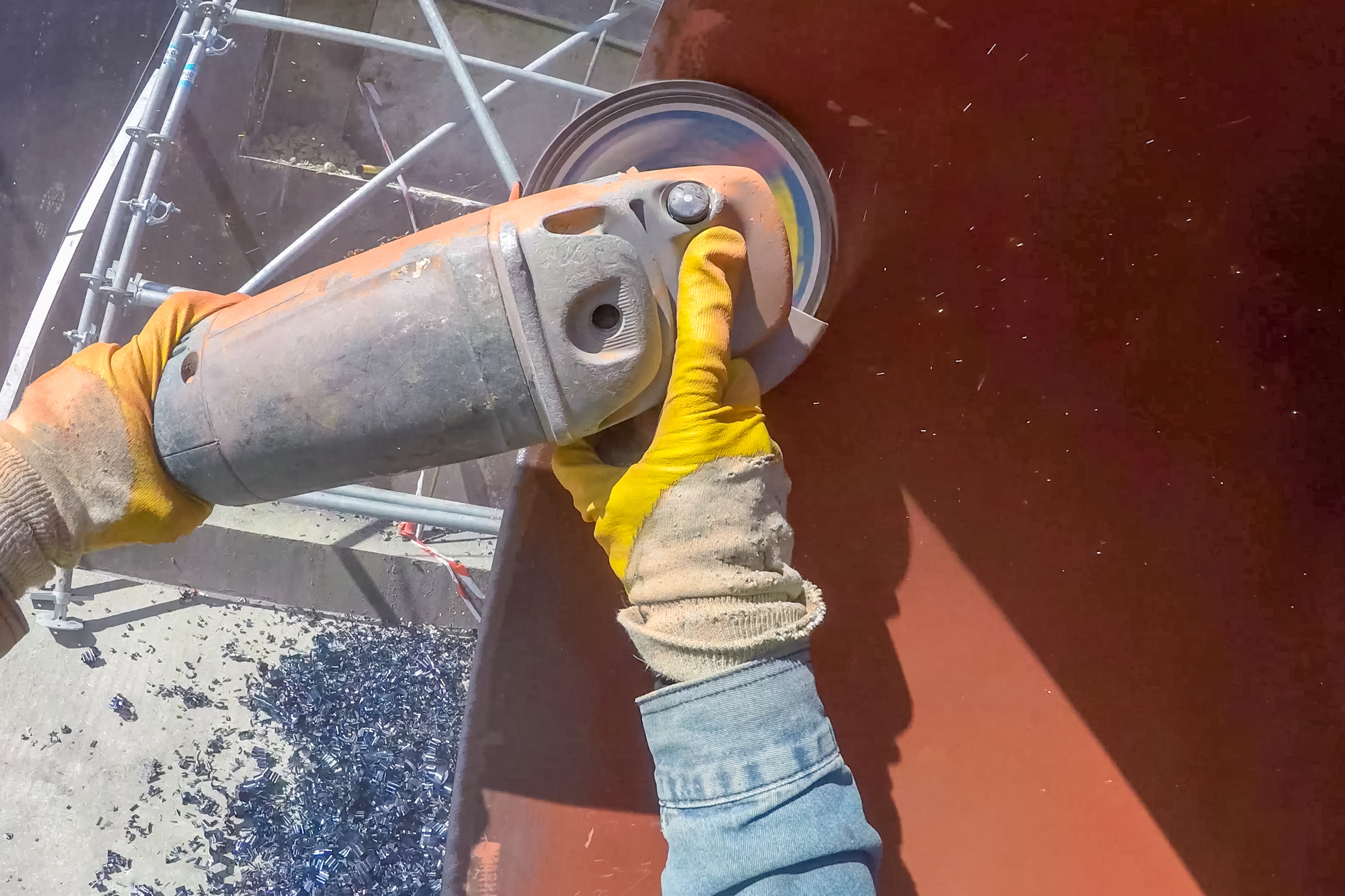 Two hands wearing yellow and white gloves using a blasting power tool on a burnt orange surface to remove the coating. There are metal pipes in the background.